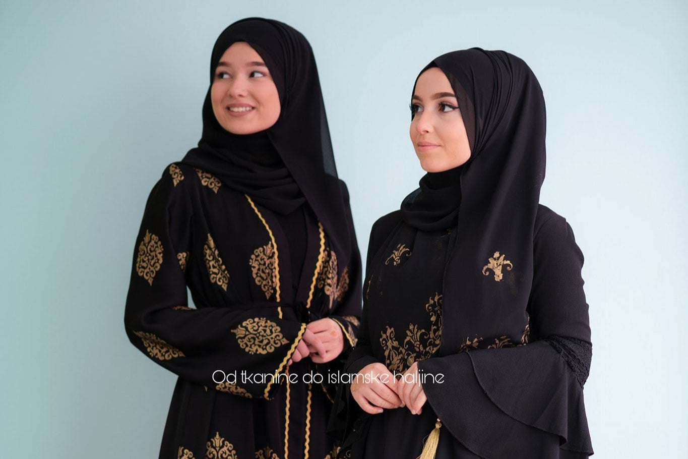 Two women wearing black abayas with gold embroidery on a light blue background